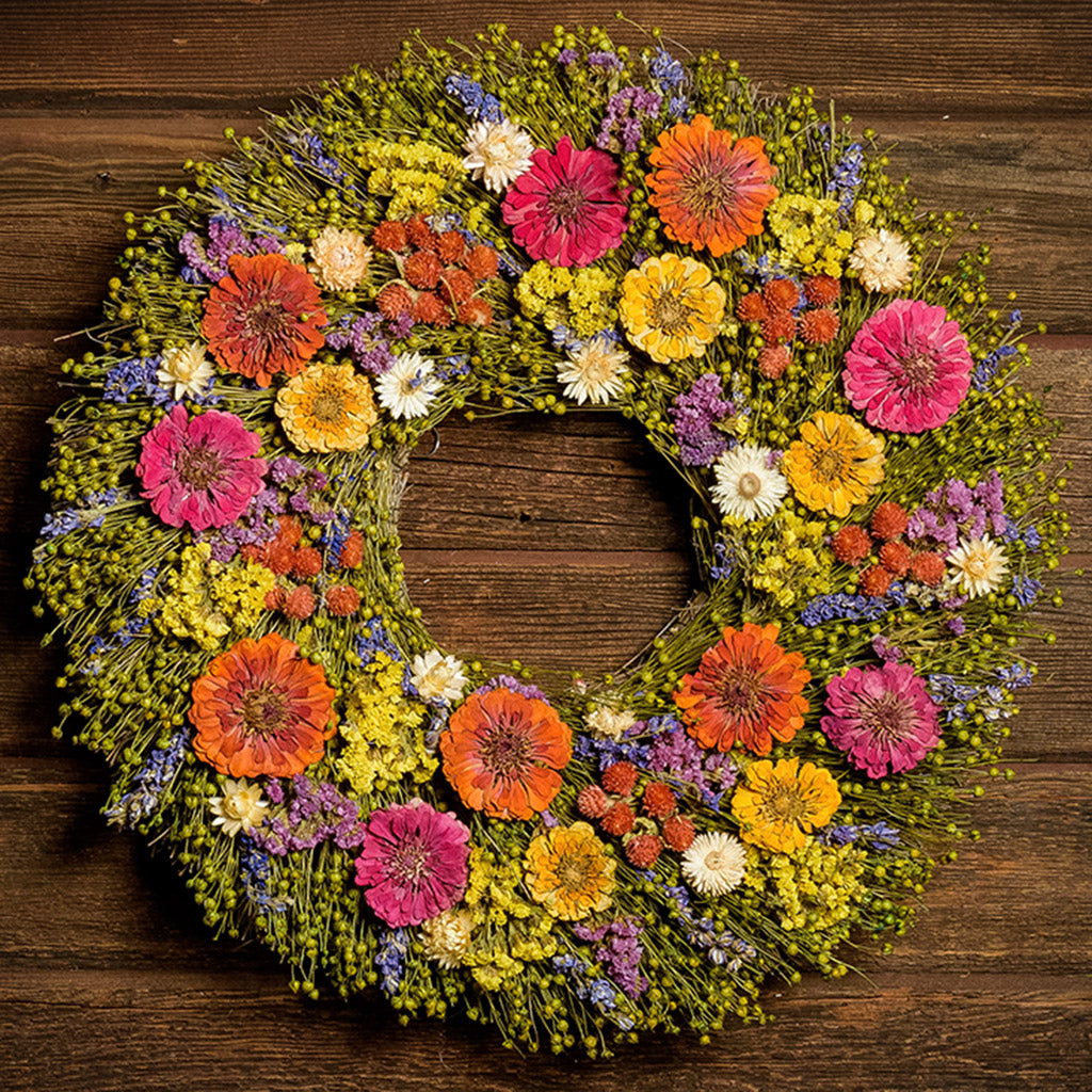 A close-up of a dried wreath made of dried yellow, pink, and orange zinnias, lavender larkspur blooms, yellow statice flowers, and white strawflowers on a base of green flax podson hung on a dark wood background.