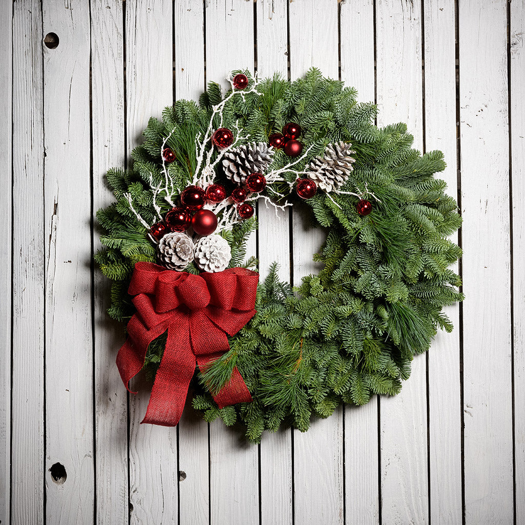 Christmas wreath with pine cones, snow-covered branches, red balls and a red burlap bow on a white wooden background