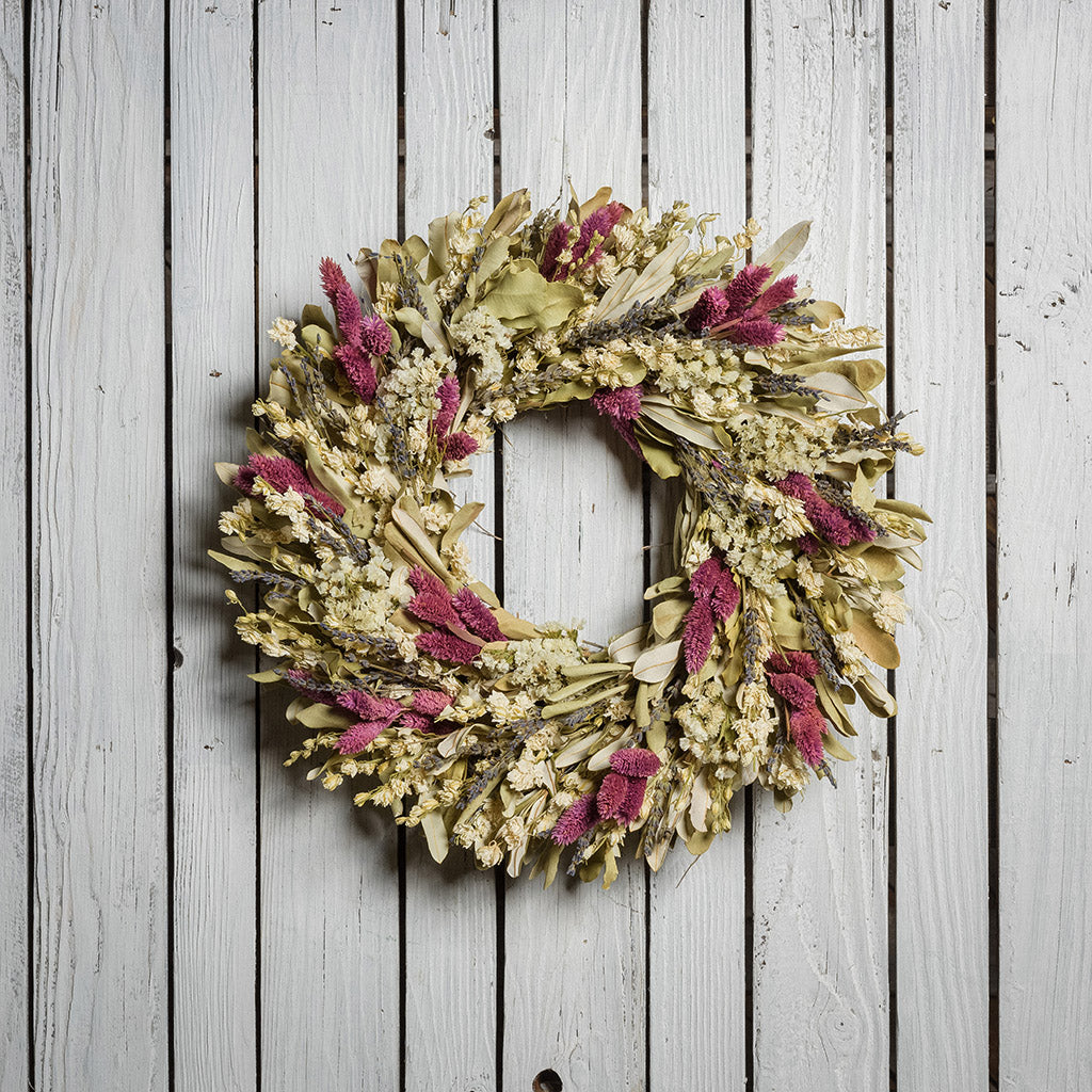 18" wreath with a blend of dried integrifolia, white larkspur, English lavender, pink phalaris, and white statice on a white wood fence background.