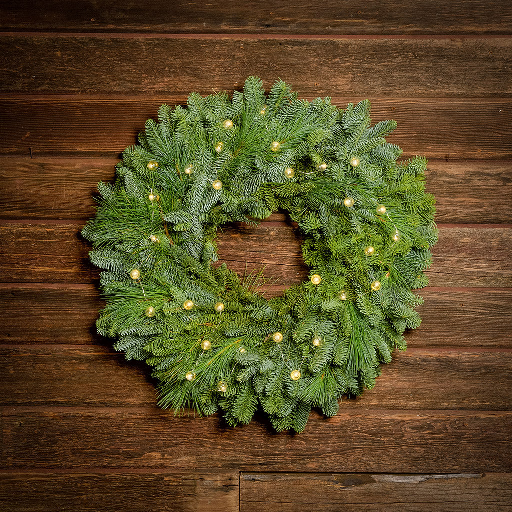 White, globe shaped, clear glass battery operated lights on a evergreen wreath hung a dark wood background.