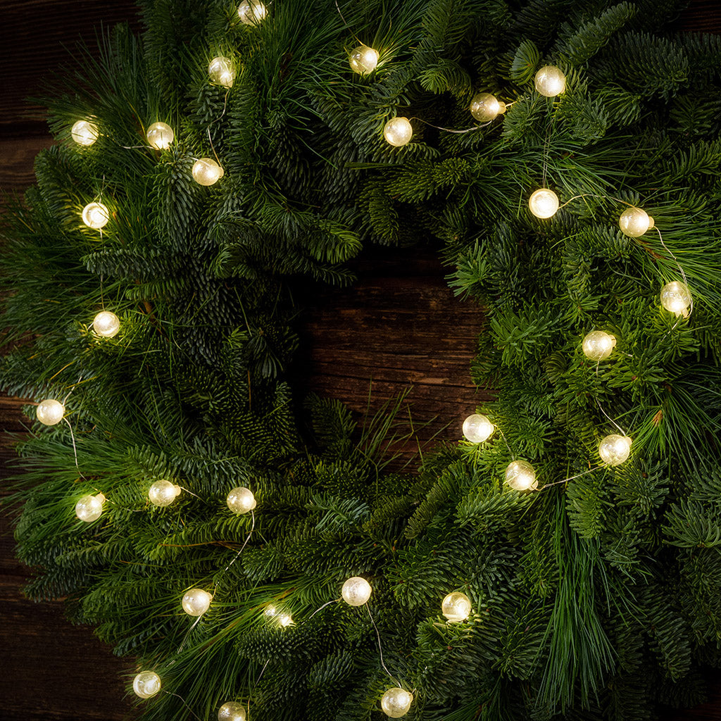 White, globe shaped, clear glass battery operated lights on a evergreen wreath hung a dark wood background.