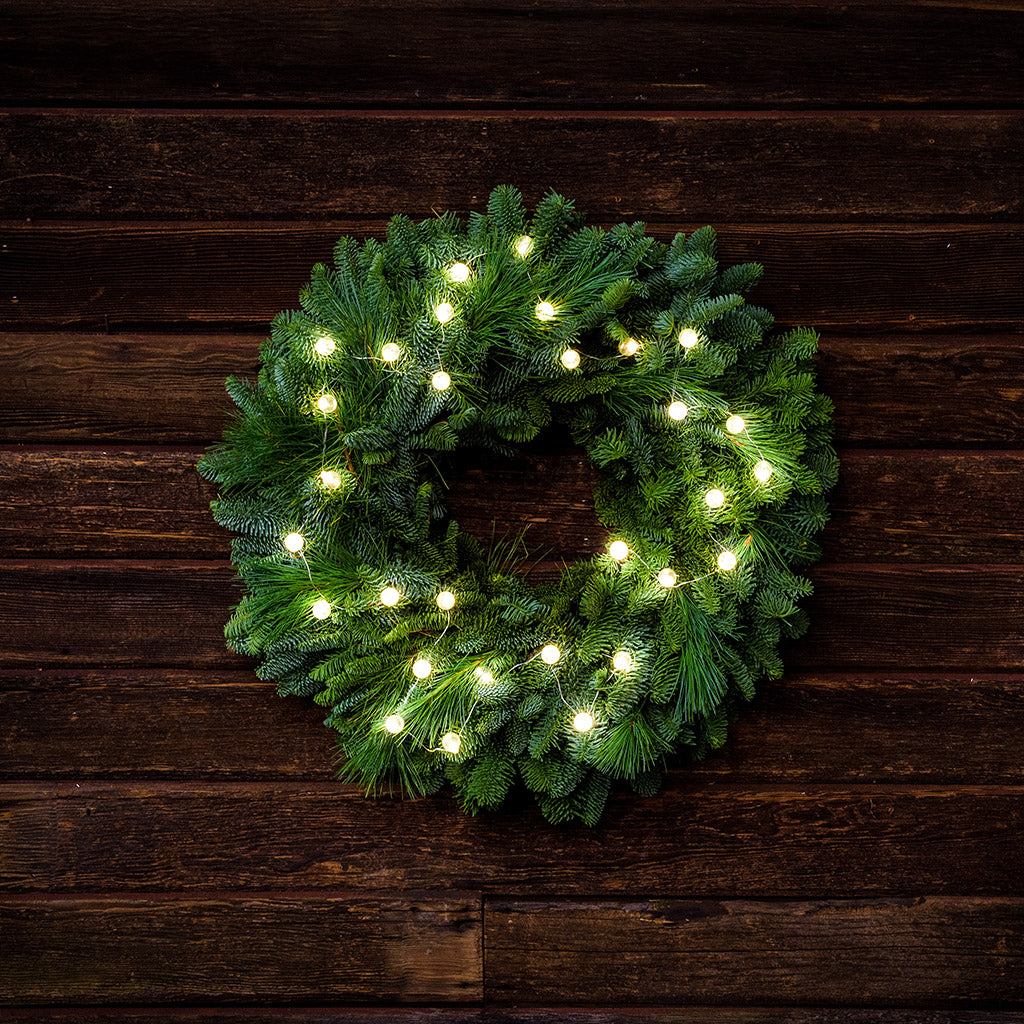 White, globe shaped, clear glass battery operated lights on a evergreen wreath hung a dark wood background.