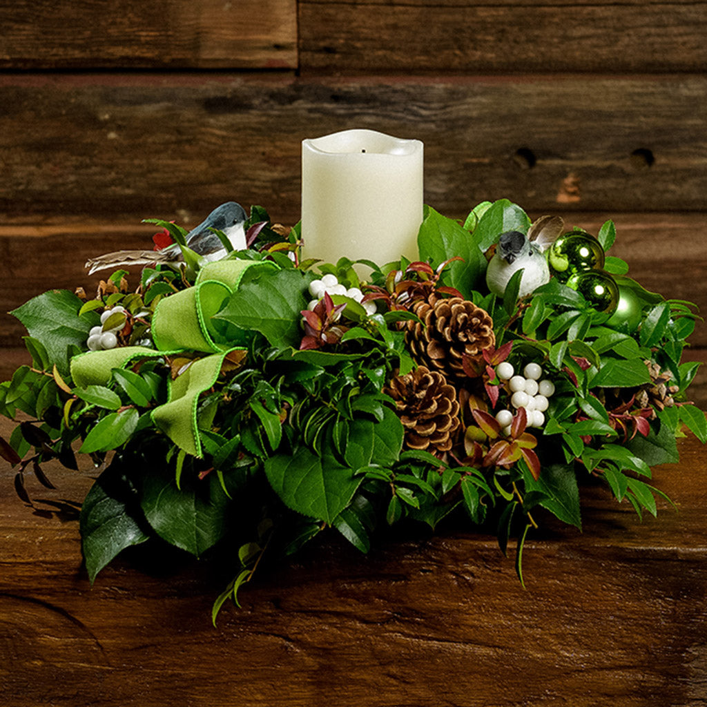 A close-up of a centerpiece made of fresh salal, green huckleberry, and red huckleberry with chickadee birds, pinecones, reindeer moss, white berry clusters, green bow tucks, and an ivory LED candle sitting on a dark wooden bench.