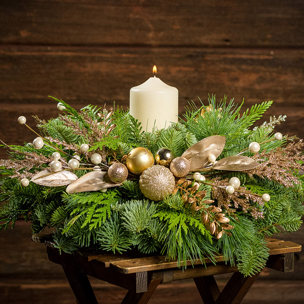 An arrangement made of noble fir, incense cedar, and white pine with gold balls, sprays of gold branches and leaves, and an ivory pillar candle with a dark wooden background.
