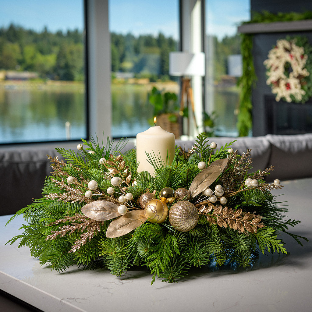 An arrangement made of noble fir, incense cedar, and white pine with gold balls, sprays of gold branches and leaves, and an ivory pillar candle sitting on a counter.