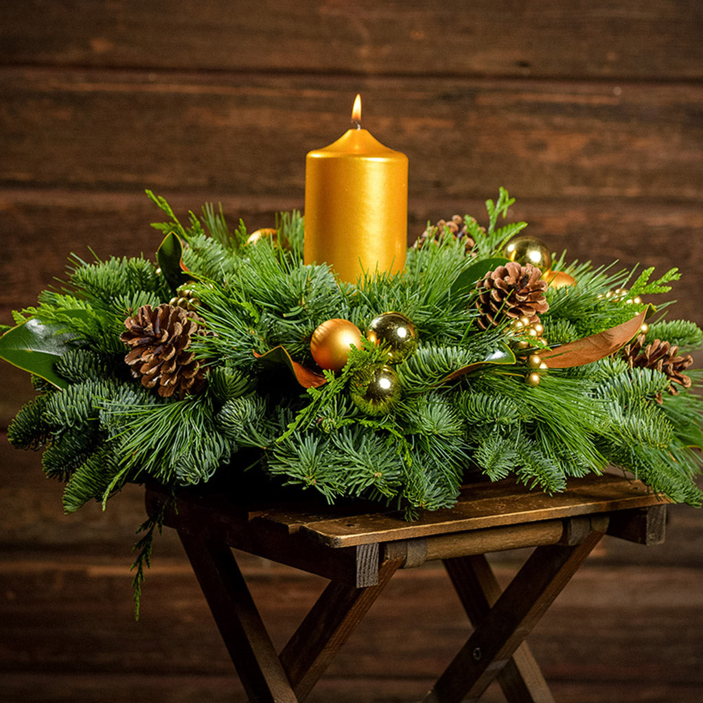 Golden magnolia centerpiece with pine cones, magnolia, gold balls and berries and a golden pillar candle with a dark wooden background.