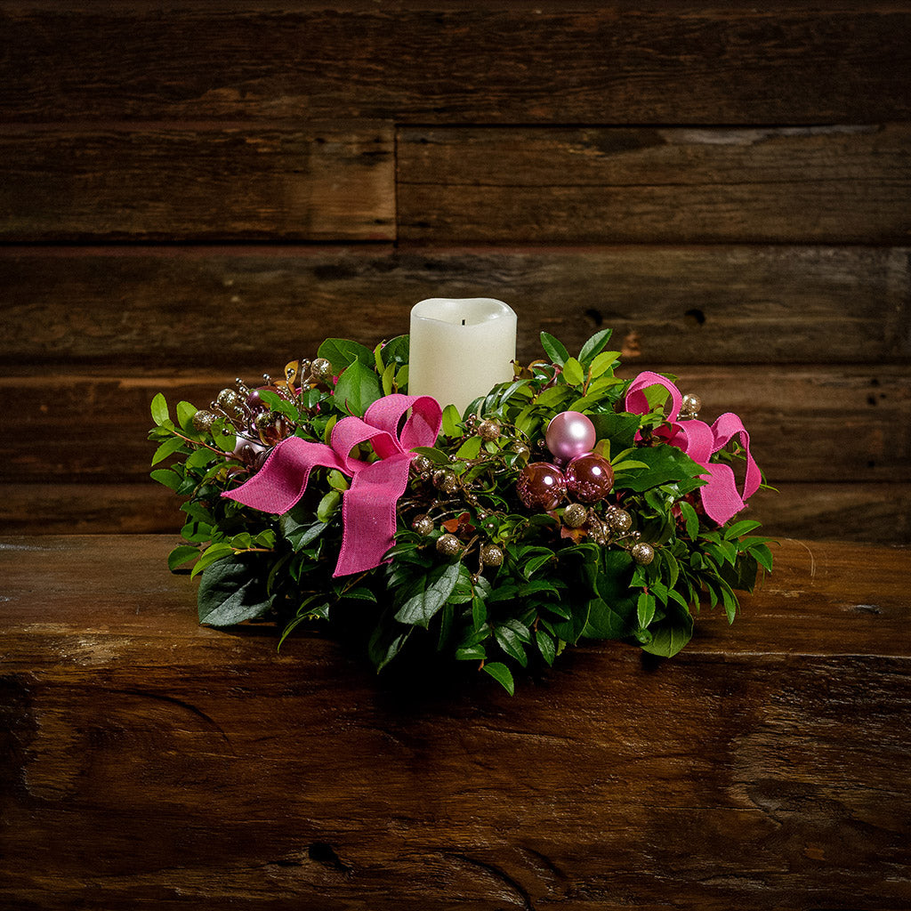 A centerpiece made of fresh salal, green huckleberry, and red huckleberry with pink ball clusters, champagne glitter berries, magenta bow tucks, and an ivory LED candle in a green plastic bowl container sitting on a dark wooden bench.