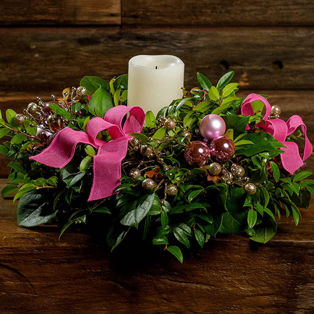 A close-up of a centerpiece made of fresh salal, green huckleberry, and red huckleberry with pink ball clusters, champagne glitter berries, magenta bow tucks, and an ivory LED candle in a green plastic bowl container sitting on a dark wooden bench.