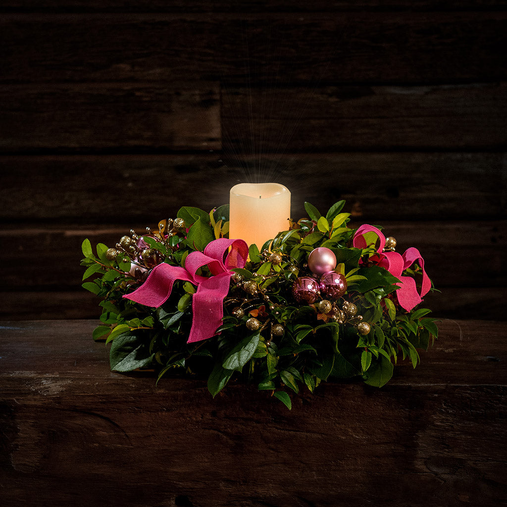 A centerpiece made of fresh salal, green huckleberry, and red huckleberry with pink ball clusters, champagne glitter berries, magenta bow tucks, and a lit ivory LED candle in a green plastic bowl container sitting on a dark wooden bench.