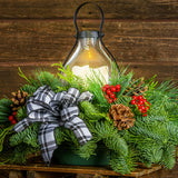 Centerpiece made of noble fir, cedar, and pine with Australian pine cones, red berry clusters, a black and white plaid bow, and an old schoolhouse-style metal LED lantern close up