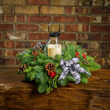 Centerpiece made of noble fir, cedar, and pine with Australian pine cones, red berry clusters, a black and white plaid bow, and an old schoolhouse-style metal LED displayed on a wooden table against a brick wall