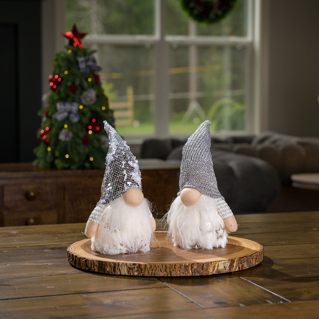 Sparkling winter light-up gnomes in fuzzy white outfits and pointy hats sitting on a wood round on a table.