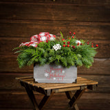 An arrangement made of noble fir, cedar, and white pine, with pinecones, red berry branches, white berry clusters, and a red and white plaid bow in a gray wood “Happy Holidays” container with a dark wooden background.