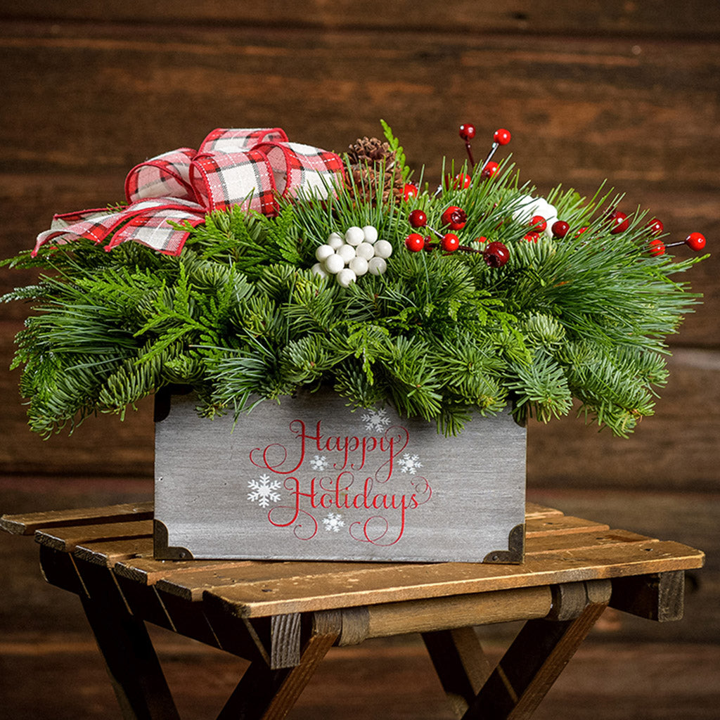 An arrangement made of noble fir, cedar, and white pine, with pinecones, red berry branches, white berry clusters, and a red and white plaid bow in a gray wood “Happy Holidays” container with a dark wooden background.