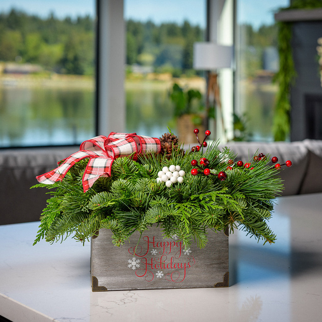 An arrangement made of noble fir, cedar, and white pine, with pinecones, red berry branches, white berry clusters, and a red and white plaid bow in a gray wood “Happy Holidays” container on a counter.