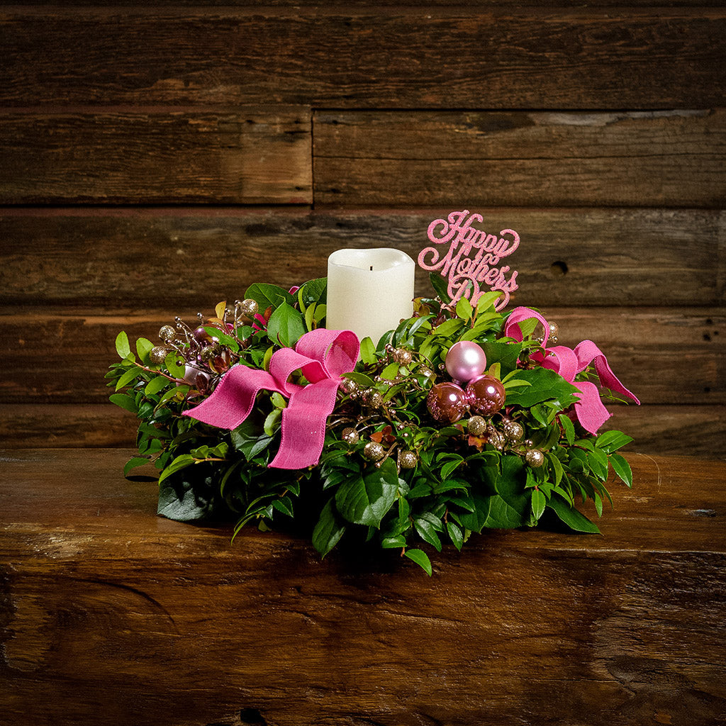 A centerpiece made of fresh salal, green huckleberry, and red huckleberry with pink ball clusters, champagne glitter berries, magenta bow tucks, a pink wooden “Happy Mother’s Day” sign, and an ivory LED candle sitting on a dark wooden bench.