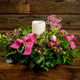 A close-up of a centerpiece made of fresh salal, green huckleberry, and red huckleberry with pink ball clusters, champagne glitter berries, magenta bow tucks, a pink wooden “Happy Mother’s Day” sign, and an ivory LED candle sitting on a dark wooden bench.