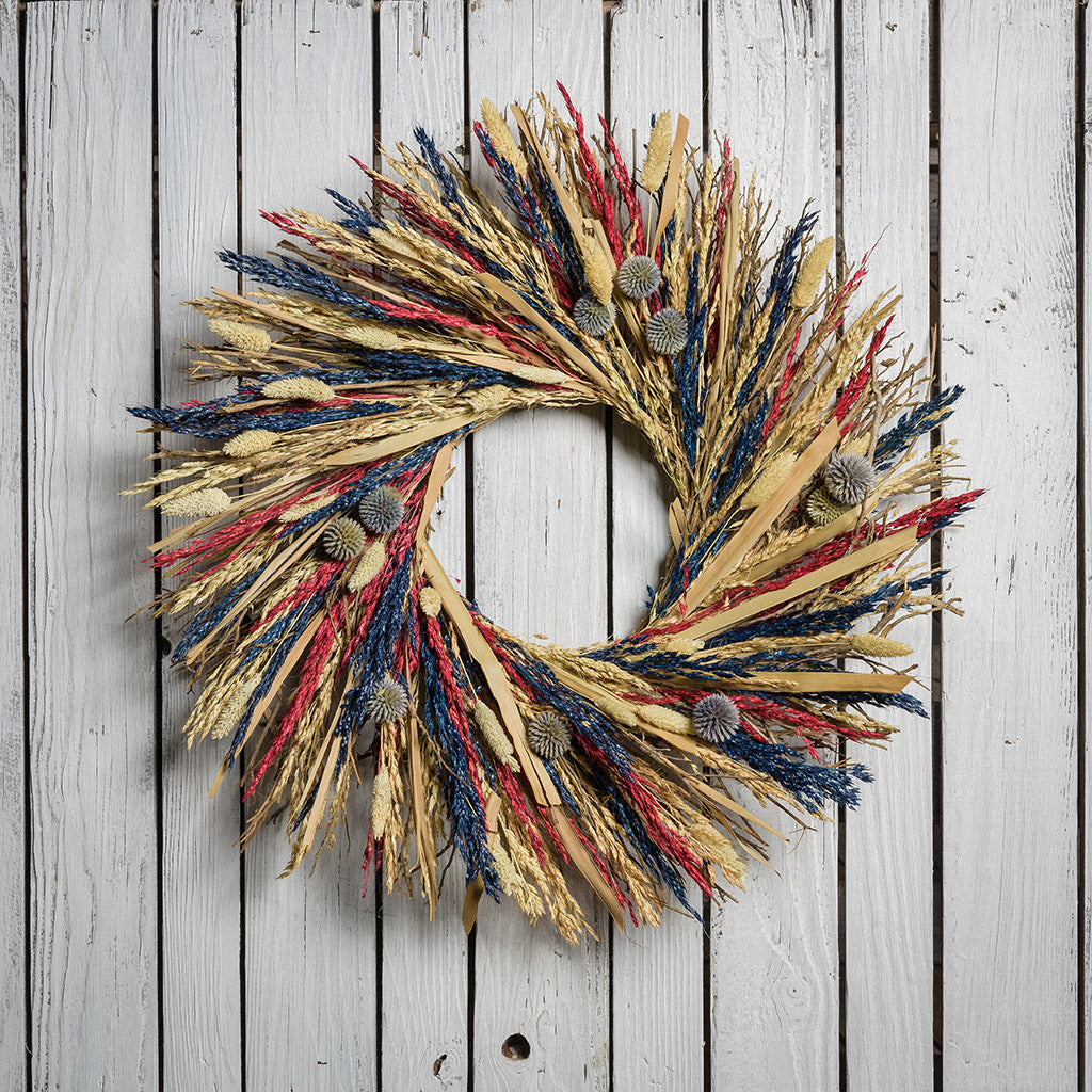 22" wreath of palm stems, blue, red and bleached sudan grass, bleached phalaris, and equinops (a small globe thistle) on a white wood fence background. 