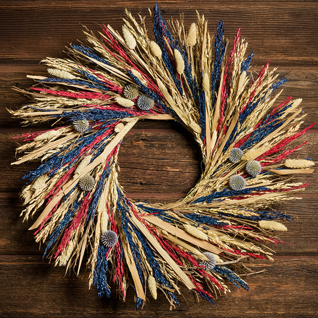 22" wreath of palm stems, blue, red and bleached sudan grass, bleached phalaris, and equinops (a small globe thistle) on a dark wood background. 
