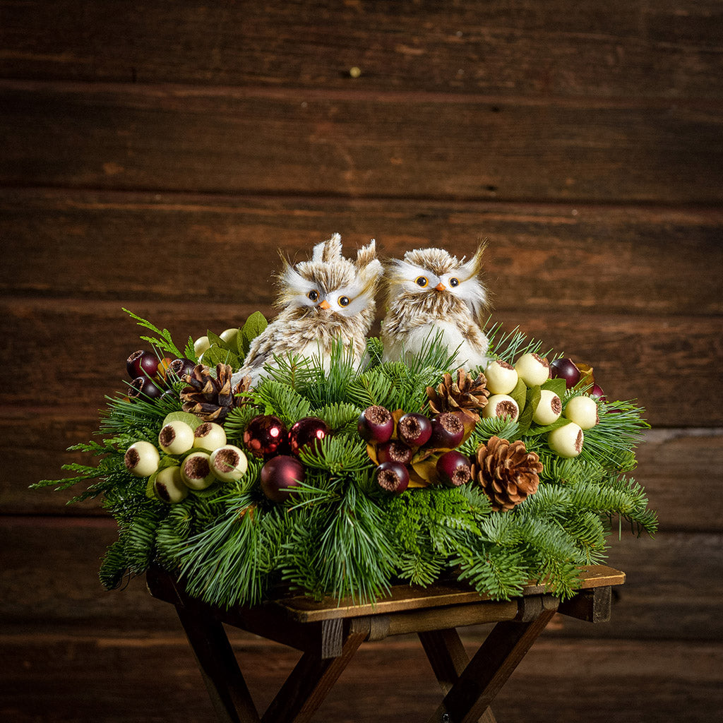 An arrangement made of noble fir, incense cedar, and white pine with burgundy ball clusters, Austrian pinecones, plum and cream berry pods with leaves, and two fluffy owls with a dark wooden background.