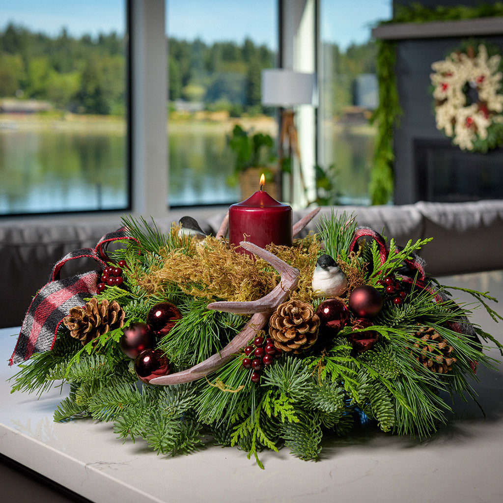 A holiday centerpiece of noble fir, white pine, incense cedar, 2 chickadee birds, 2 red and black plaid bows, 4 burgundy ball clusters, 6 Australian pinecones, 6 burgundy berry clusters, moss, 2 faux antlers, and a burgundy pillar candle sitting on a counter.