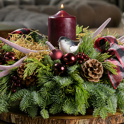 A holiday centerpiece of noble fir, white pine, incense cedar, 2 chickadee birds, 2 red and black plaid bows, 4 burgundy ball clusters, 6 Australian pinecones, 6 burgundy berry clusters, moss, 2 faux antlers, and a burgundy pillar candle sitting on a wood round. 