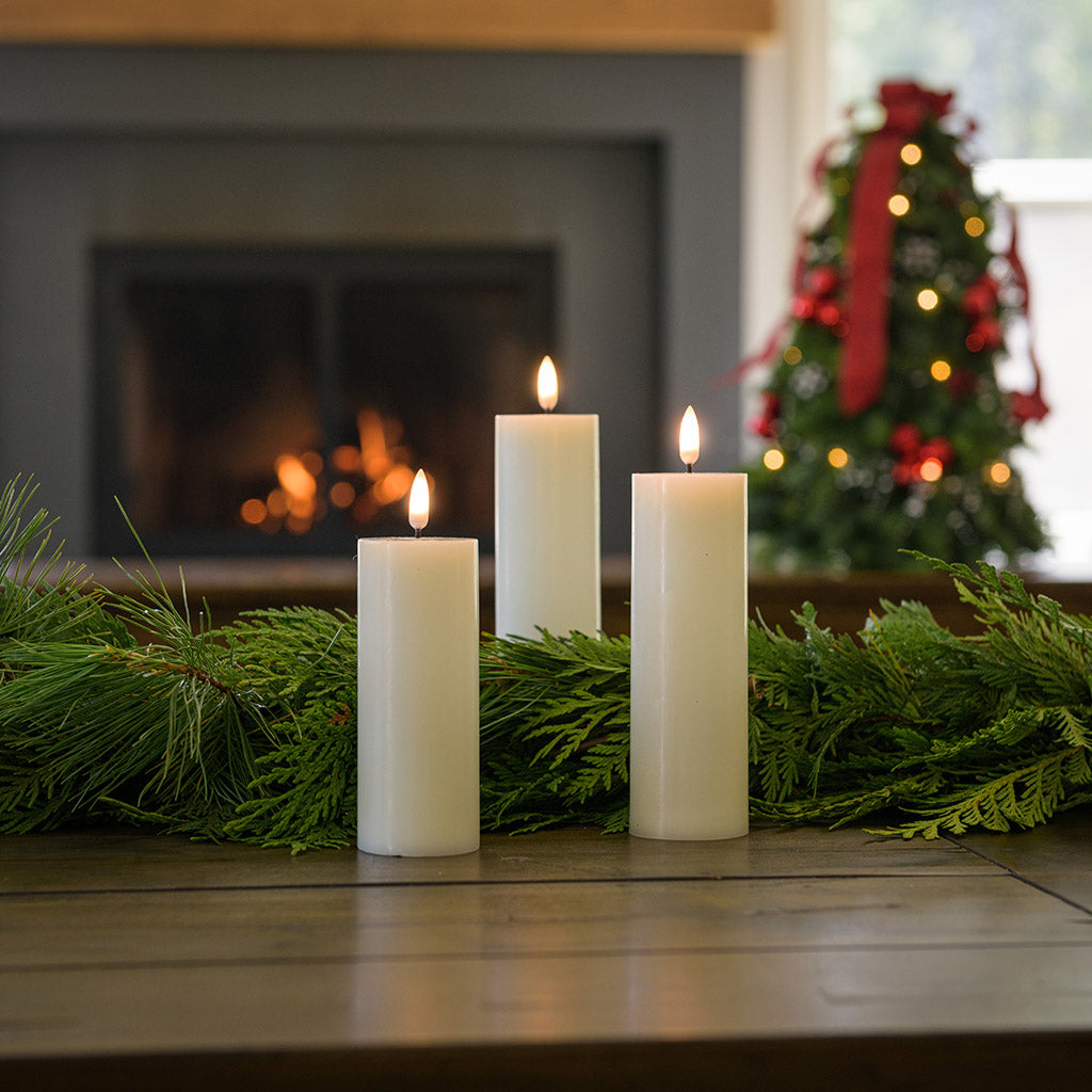 Set of 3 ivory LED candles with a 360 degree flame paired with garland on a wood table.
