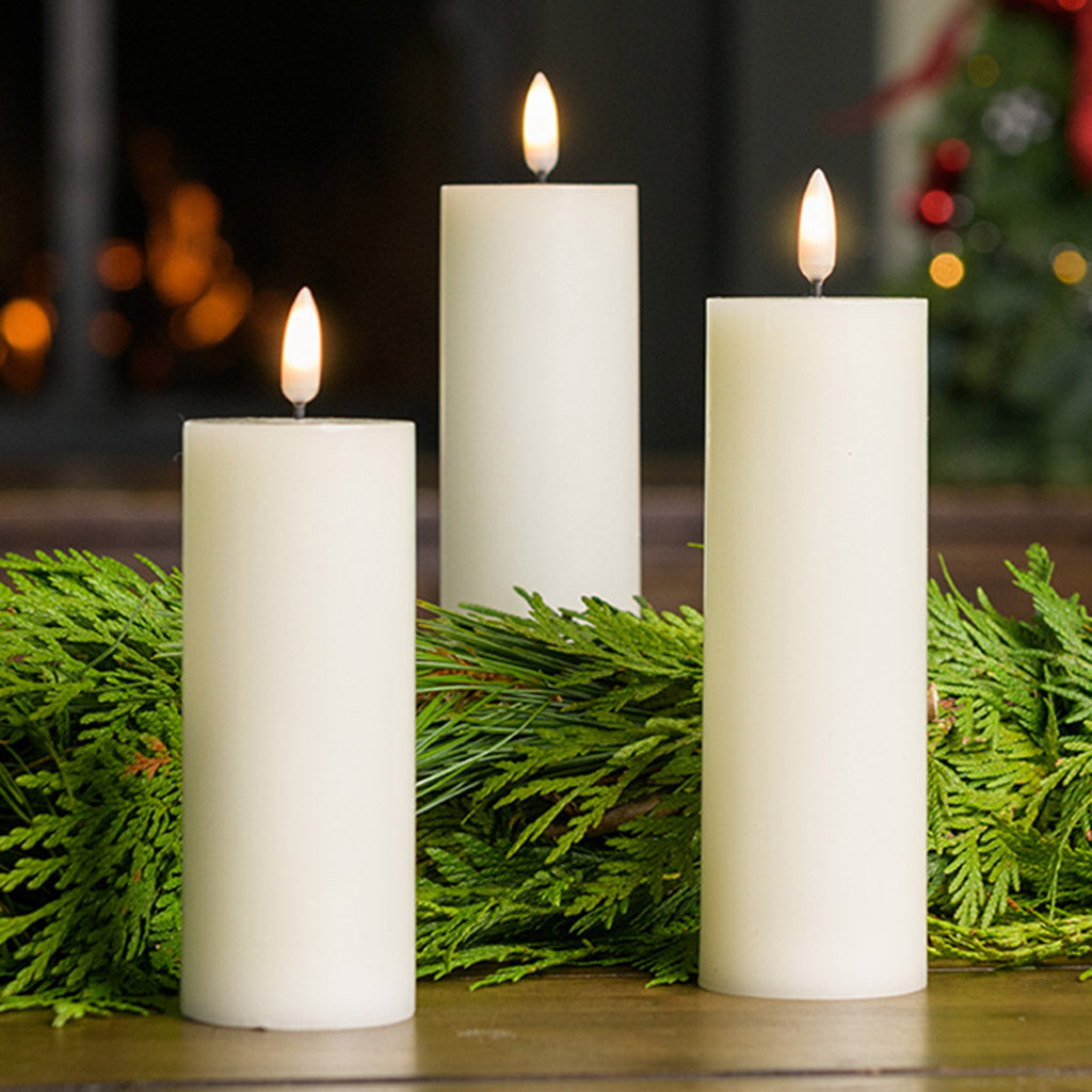 Set of 3 ivory LED candles with a 360 degree flame paired with garland on a wood table.