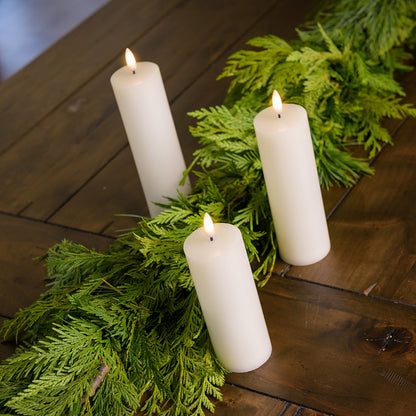 Set of 3 ivory LED candles with a 360 degree flame paired with garland on a wood table.