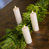 Set of 3 ivory LED candles with a 360 degree flame paired with garland on a wood table.