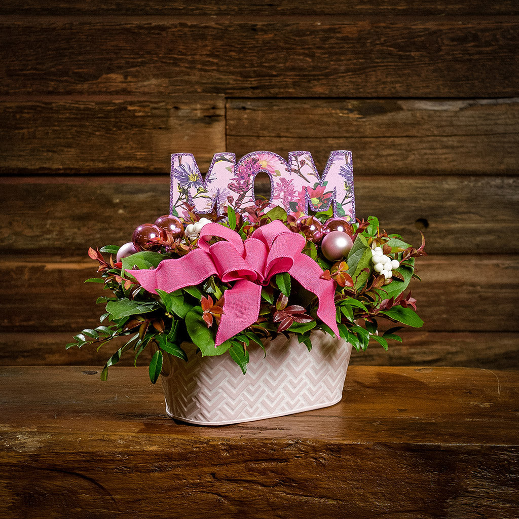 A centerpiece made of fresh salal, green huckleberry, red huckleberry, sweet huckleberry with pink ball clusters, white berries, a magenta pink bow, and a purple wood “MOM” sign in a pink and white metal container on a dark wooden bench.