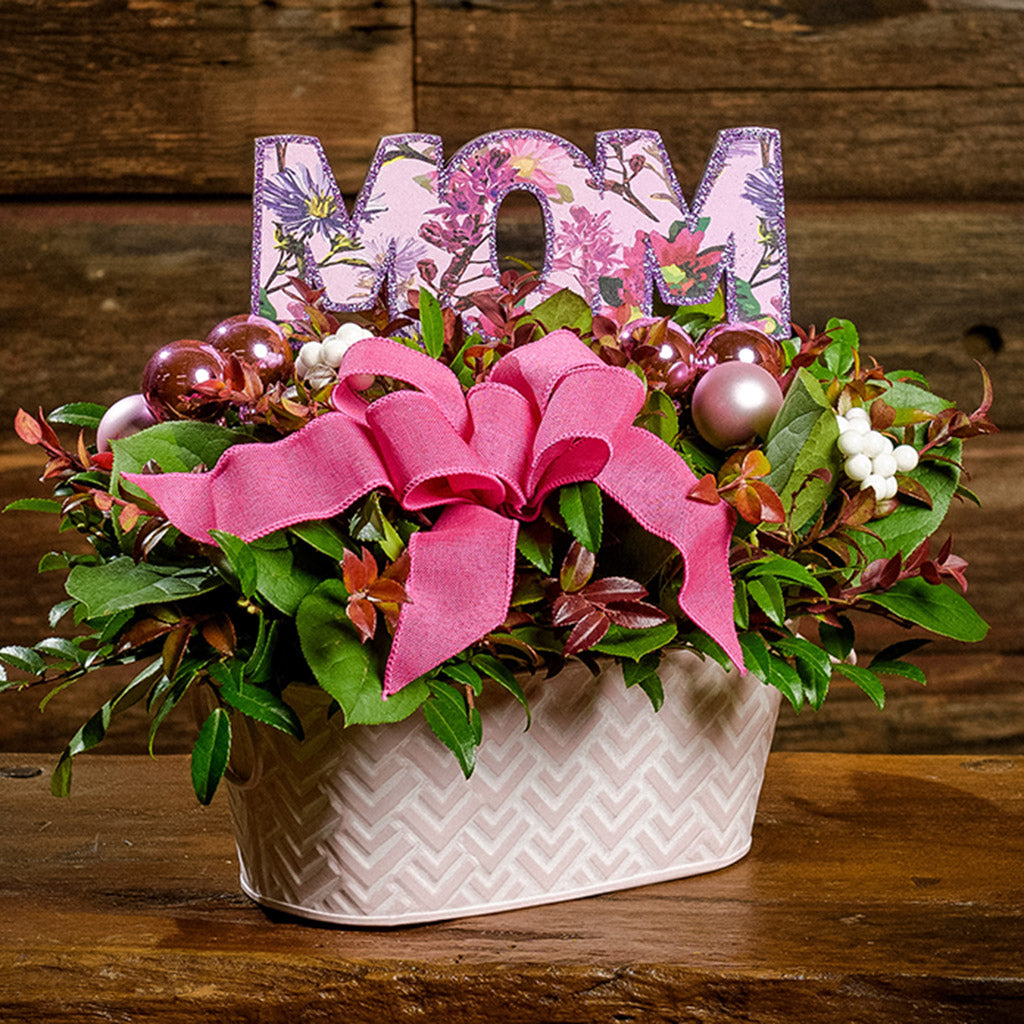 A close-up of a centerpiece made of fresh salal, green huckleberry, red huckleberry, sweet huckleberry with pink ball clusters, white berries, a magenta pink bow, and a purple wood “MOM” sign in a pink and white metal container on a dark wooden bench.