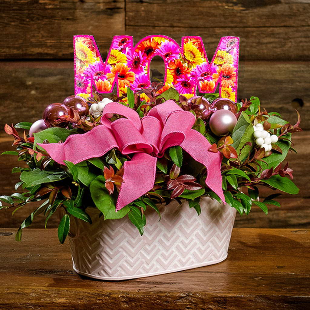 A close-up of a centerpiece made of fresh salal, green huckleberry, red huckleberry, sweet huckleberry with pink ball clusters, white berries, a magenta pink bow, and a wood “MOM” sign in a pink and white metal container.