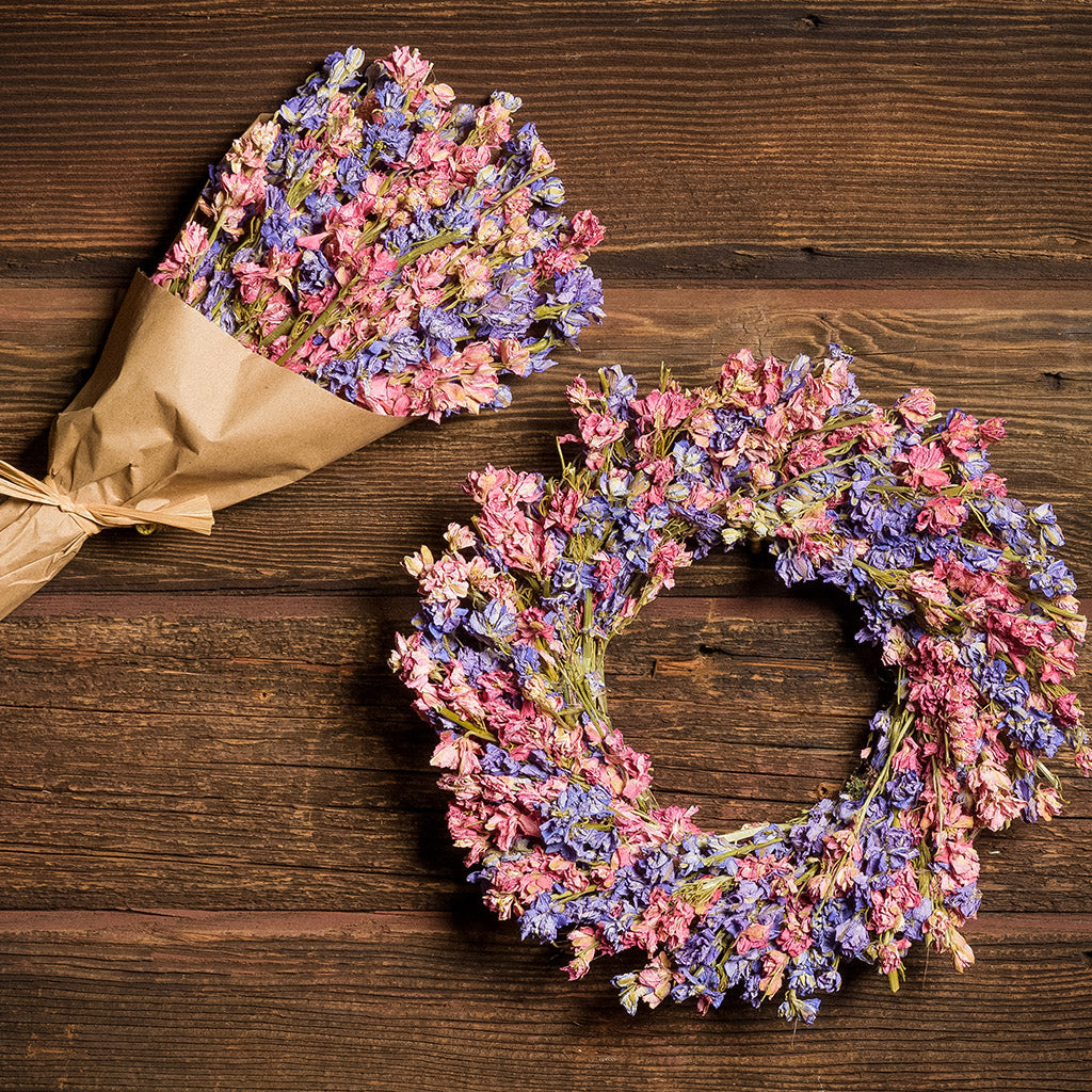 10” dried larkspur wreath with mixed pink and lavender larkspur is accompanied by a matching 11” bouquet of dried larkspur wrapped in kraft paper on a dark wood background. 