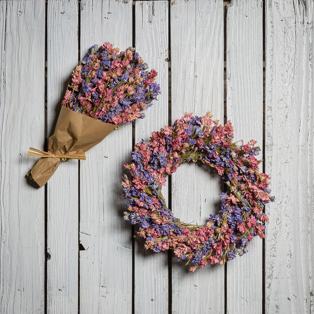 10” dried larkspur wreath with mixed pink and lavender larkspur is accompanied by a matching 11” bouquet of dried larkspur wrapped in kraft paper on a white wood fence background.