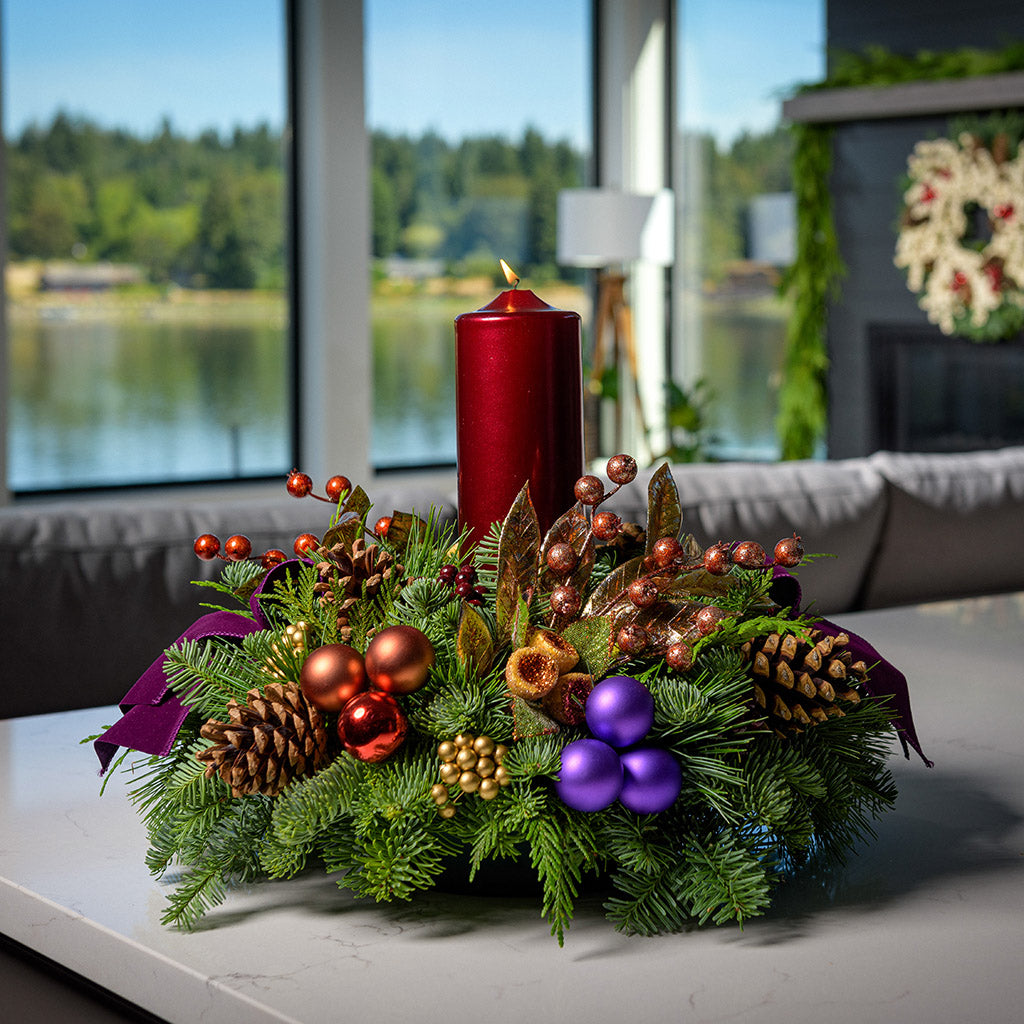 An arrangement made of noble fir, incense cedar, and white pine with Austrian pinecones, gold and burgundy berry clusters, purple and copper ball clusters, copper berry and leaf branches, purple velvet bow tucks, and a tall pomegranate pillar candle sitting on a counter.
