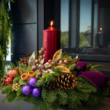 An arrangement made of noble fir, incense cedar, and white pine with Austrian pinecones, gold and burgundy berry clusters, purple and copper ball clusters, copper berry and leaf branches, purple velvet bow tucks, and a tall pomegranate pillar candle sitting next to a fireplace.