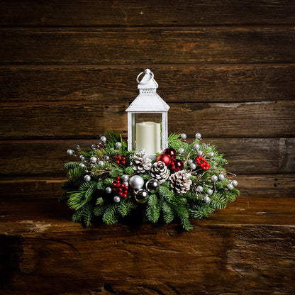 A holiday centerpiece of noble fir, white pine, incense cedar, silver glittery bead decorations, 2 red ball clusters, 2 silver ball clusters, 6 red berry clusters, 6 Australian pinecones with silver glitter, and an LED white lantern with a wood background. 