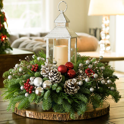 A holiday centerpiece of noble fir, white pine, incense cedar, silver glittery bead decorations, 2 red ball clusters, 2 silver ball clusters, 6 red berry clusters, 6 Australian pinecones with silver glitter, and an LED white lantern sitting on a wood round on a table up close.