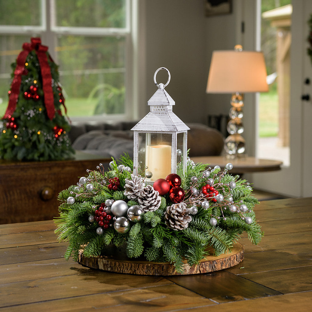 A holiday centerpiece of noble fir, white pine, incense cedar, silver glittery bead decorations, 2 red ball clusters, 2 silver ball clusters, 6 red berry clusters, 6 Australian pinecones with silver glitter, and an LED white lantern sitting on a wood round on a table. 