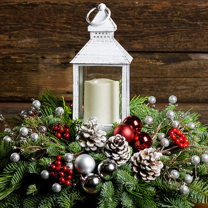 A holiday centerpiece of noble fir, white pine, incense cedar, silver glittery bead decorations, 2 red ball clusters, 2 silver ball clusters, 6 red berry clusters, 6 Australian pinecones with silver glitter, and an LED white lantern with wood background. 