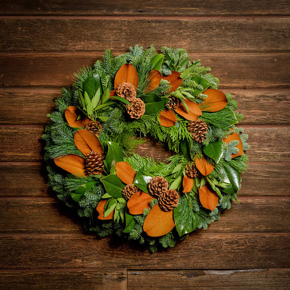 Holiday wreath made of noble fir, cedar, and pine with bay and magnolia leaves, ponderosa pine cones, and Australian pine cones with a dark wood backgound.