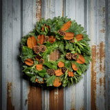 Holiday wreath made of noble fir, cedar, and pine with bay and magnolia leaves, ponderosa pine cones, and Australian pine cones on a rustic metal wall.