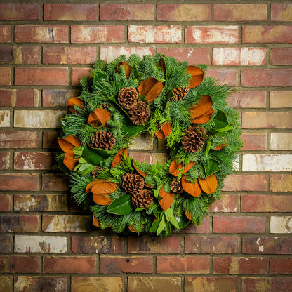 Holiday wreath made of noble fir, cedar, and pine with bay and magnolia leaves, ponderosa pine cones, and Australian pine cones hanging on a brick wall