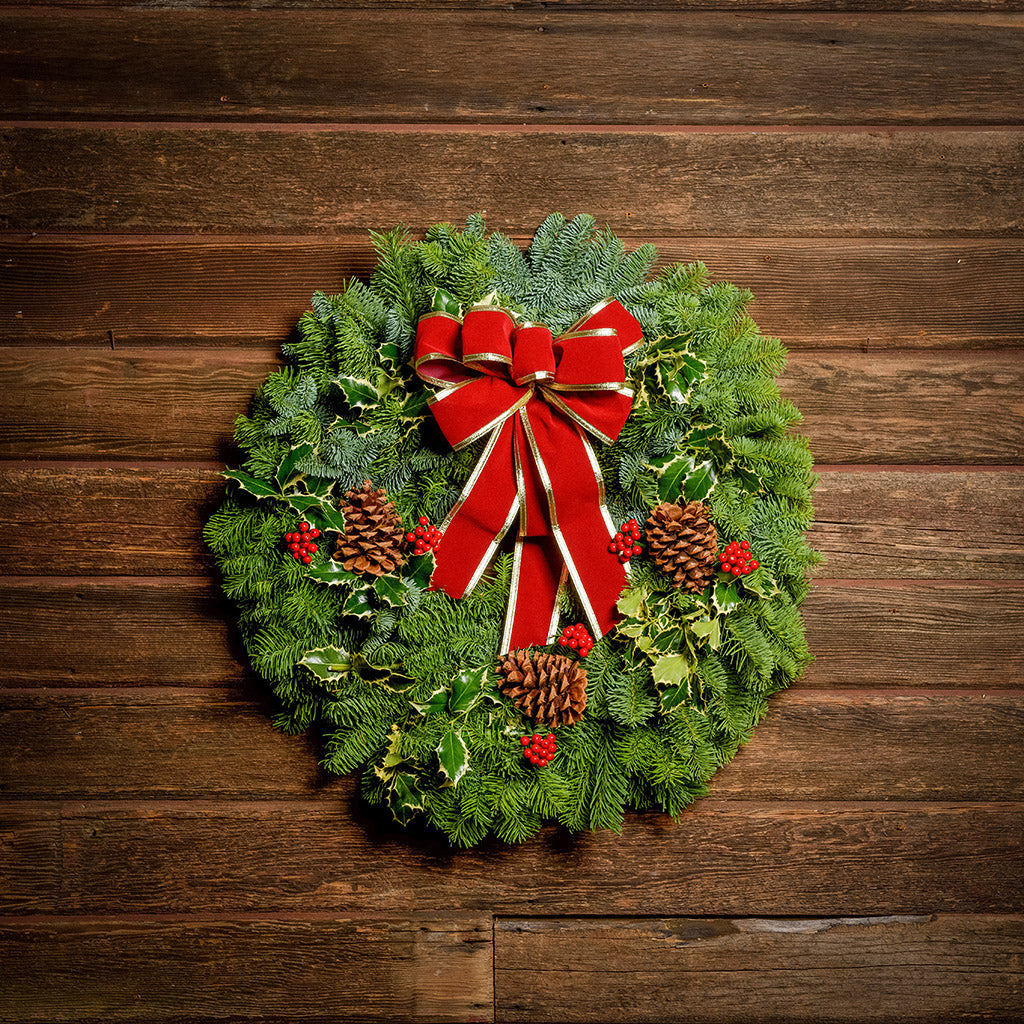 Christmas wreath made of variegated holly, noble fir, incense cedar and juniper with ponderosa pine cones, faux red berry clusters and a red velveteen with gold back bow hanging on a wooden wall