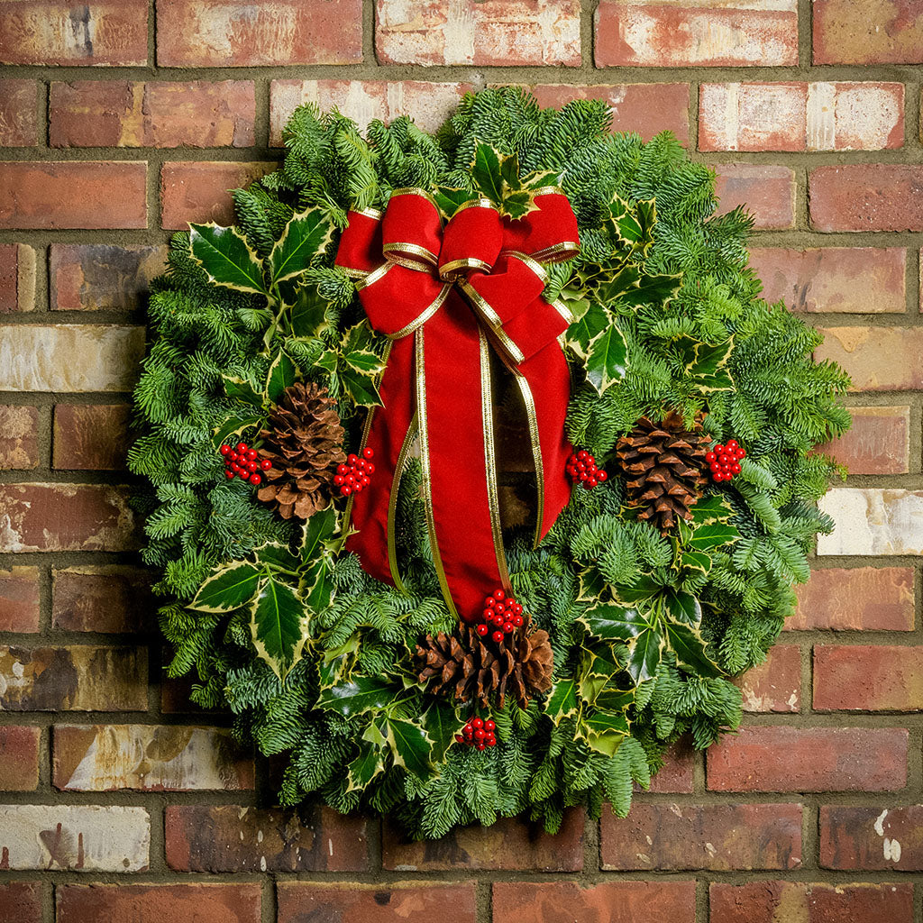Christmas wreath made of variegated holly, noble fir, incense cedar and juniper with ponderosa pine cones, faux red berry clusters and a red velveteen with gold back bow hanging on a brick wall