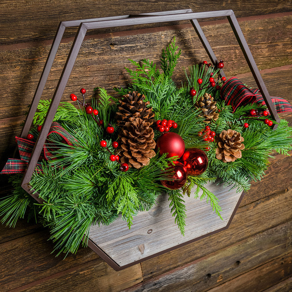 Centerpiece with noble fir, cedar, and pine with 2 ponderosa pine cones, 2 Australian pine cones, 1 red ball cluster, 4 red berry branches, 2 red plaid bow tucks, and a hexagon metal and wooden container mounted on a wooden wall