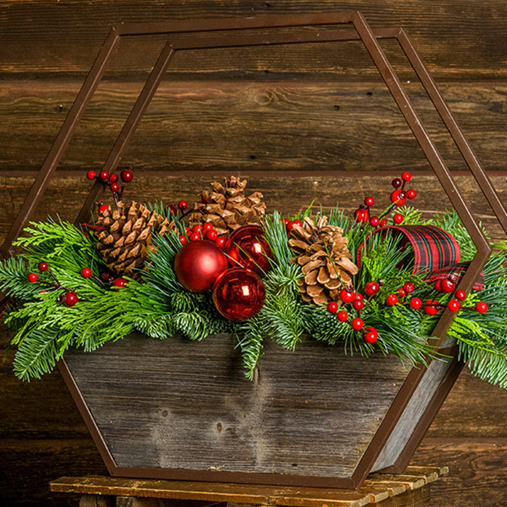 Centerpiece with noble fir, cedar, and pine with 2 ponderosa pine cones, 2 Australian pine cones, 1 red ball cluster, 4 red berry branches, 2 red plaid bow tucks, and a hexagon metal and wooden container on a wooden bench