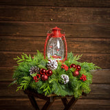 An arrangement of noble fir, incense cedar, and white pine with frosted pinecones, red berry clusters, red ball clusters, and a wintery scene in a red globe lantern with a dark wooden background.