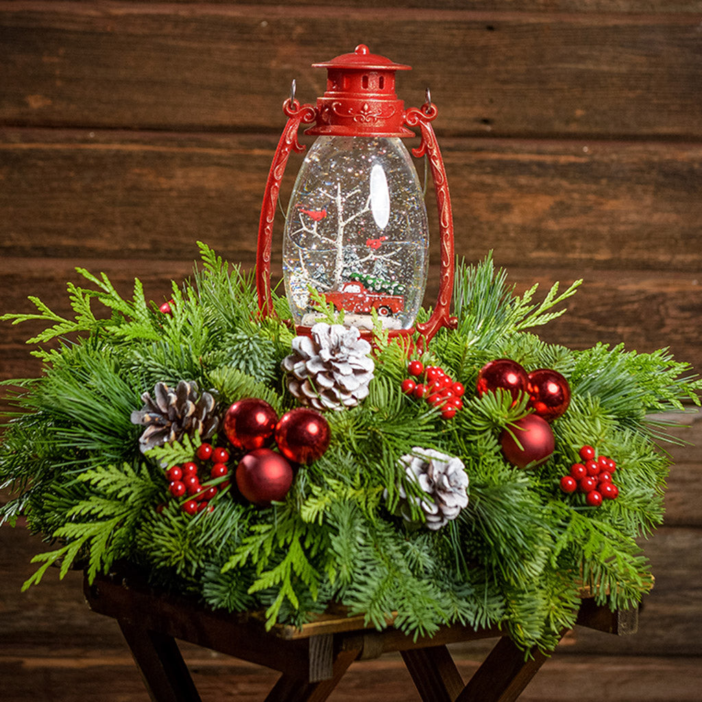 An arrangement of noble fir, incense cedar, and white pine with frosted pinecones, red berry clusters, red ball clusters, and a wintery scene in a red globe lantern with a dark wooden background.